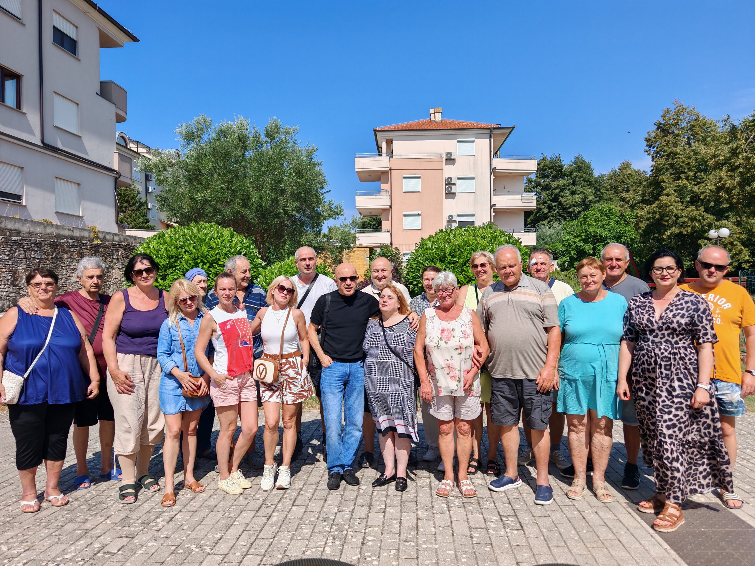 Na fotografiji je veća skupina sudionika edukacijsko-rehabilitacijskog kampa Udruge slijepih USKA, okupljena na otvorenom za zajedničku fotografiju. Sudionici stoje u nekoliko redova, nasmijani i opušteni, a među njima su osobe različite dobi. Odjeveni su u ljetnu odjeću, što odgovara ljetnom boravku u Premanturi. U pozadini se vide stambene zgrade, zelenilo i vedro plavo nebo, dok je cijela skupina obasjana sunčevom svjetlošću. Fotografija dočarava zajedništvo, druženje i dobro raspoloženje sudionika kampa te lijepo prikazuje atmosferu zajedničkog boravka i aktivnosti.