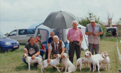 Skupina muškaraca sa psima na travnatoj površini ispred šatora i parkiranih vozila.