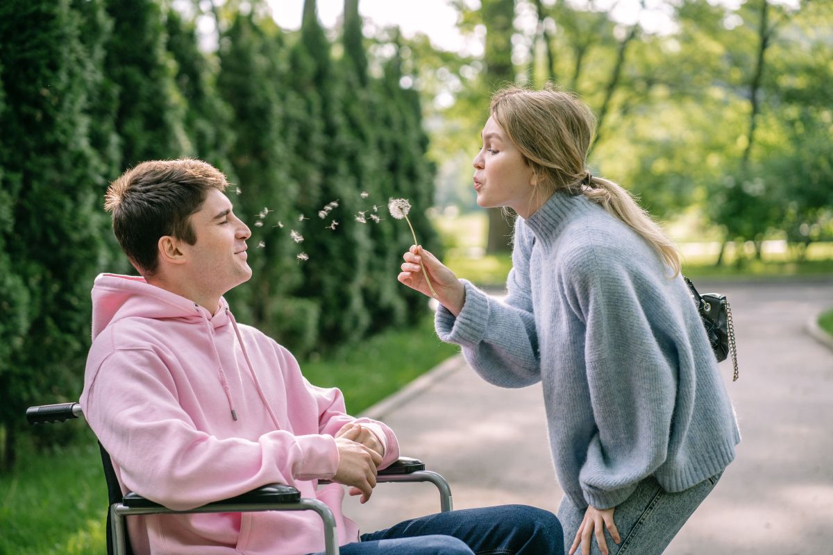 Žena puše u maslačak pokraj muškarca u invalidskim kolicima u parku.
