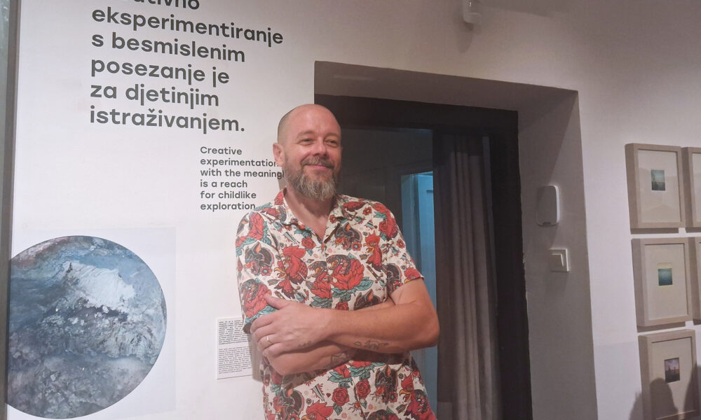 Muškarac u šarenoj košulji stoji u galerijskom prostoru okružen umjetničkim radovima.