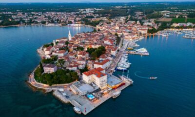 Fotografija prikazuje Poreč snimljen iz zraka, u panoramskom pogledu iznad mora. U prvom planu ističe se povijesna gradska jezgra smještena na poluotoku, okružena Jadranskim morem. U središtu fotografije vidljive su brojne kuće i zgrade s karakterističnim crvenim krovovima, uske ulice te prepoznatljivi zvonik. Uz obalu se protežu uređena šetnica i marina s brojnim jedrilicama, brodicama i jahtama. Posebno se ističe pogled na staru gradsku jezgru koja se pruža između dviju morskih površina. U pozadini se vidi šire urbano područje Poreča, okruženo zelenilom, šumama, poljoprivrednim površinama i manjim naseljima. Fotografija je snimljena u večernjim satima, pa se uz obalu već vide upaljena svjetla restorana, hotela i drugih objekata. Plavo more, povijesna arhitektura, marina i zeleni krajolik zajedno stvaraju prepoznatljivu sliku Poreča kao mediteranskog turističkog grada bogate kulturne baštine i razvijenog pomorskog turizma.