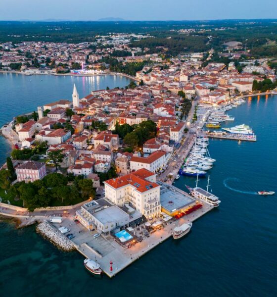 Fotografija prikazuje Poreč snimljen iz zraka, u panoramskom pogledu iznad mora. U prvom planu ističe se povijesna gradska jezgra smještena na poluotoku, okružena Jadranskim morem. U središtu fotografije vidljive su brojne kuće i zgrade s karakterističnim crvenim krovovima, uske ulice te prepoznatljivi zvonik. Uz obalu se protežu uređena šetnica i marina s brojnim jedrilicama, brodicama i jahtama. Posebno se ističe pogled na staru gradsku jezgru koja se pruža između dviju morskih površina. U pozadini se vidi šire urbano područje Poreča, okruženo zelenilom, šumama, poljoprivrednim površinama i manjim naseljima. Fotografija je snimljena u večernjim satima, pa se uz obalu već vide upaljena svjetla restorana, hotela i drugih objekata. Plavo more, povijesna arhitektura, marina i zeleni krajolik zajedno stvaraju prepoznatljivu sliku Poreča kao mediteranskog turističkog grada bogate kulturne baštine i razvijenog pomorskog turizma.
