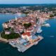 Fotografija prikazuje Poreč snimljen iz zraka, u panoramskom pogledu iznad mora. U prvom planu ističe se povijesna gradska jezgra smještena na poluotoku, okružena Jadranskim morem. U središtu fotografije vidljive su brojne kuće i zgrade s karakterističnim crvenim krovovima, uske ulice te prepoznatljivi zvonik. Uz obalu se protežu uređena šetnica i marina s brojnim jedrilicama, brodicama i jahtama. Posebno se ističe pogled na staru gradsku jezgru koja se pruža između dviju morskih površina. U pozadini se vidi šire urbano područje Poreča, okruženo zelenilom, šumama, poljoprivrednim površinama i manjim naseljima. Fotografija je snimljena u večernjim satima, pa se uz obalu već vide upaljena svjetla restorana, hotela i drugih objekata. Plavo more, povijesna arhitektura, marina i zeleni krajolik zajedno stvaraju prepoznatljivu sliku Poreča kao mediteranskog turističkog grada bogate kulturne baštine i razvijenog pomorskog turizma.