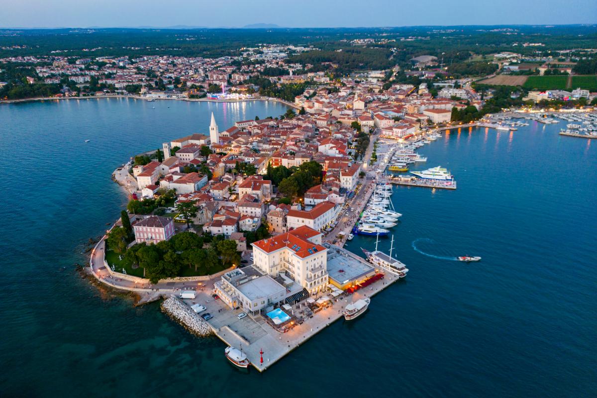 Fotografija prikazuje Poreč snimljen iz zraka, u panoramskom pogledu iznad mora. U prvom planu ističe se povijesna gradska jezgra smještena na poluotoku, okružena Jadranskim morem. U središtu fotografije vidljive su brojne kuće i zgrade s karakterističnim crvenim krovovima, uske ulice te prepoznatljivi zvonik. Uz obalu se protežu uređena šetnica i marina s brojnim jedrilicama, brodicama i jahtama. Posebno se ističe pogled na staru gradsku jezgru koja se pruža između dviju morskih površina. U pozadini se vidi šire urbano područje Poreča, okruženo zelenilom, šumama, poljoprivrednim površinama i manjim naseljima. Fotografija je snimljena u večernjim satima, pa se uz obalu već vide upaljena svjetla restorana, hotela i drugih objekata. Plavo more, povijesna arhitektura, marina i zeleni krajolik zajedno stvaraju prepoznatljivu sliku Poreča kao mediteranskog turističkog grada bogate kulturne baštine i razvijenog pomorskog turizma.