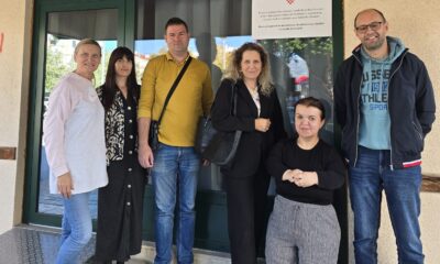 Na fotografiji je šest osoba okupljenih ispred ulaza u zgradu, pozirajući za zajedničku fotografiju. Skupinu čine predstavnici partnerskih organizacija i korisnica projekta, a iza njih na staklenim vratima istaknut je plakat s grbom Republike Hrvatske i informacijom o Programu prekogranične suradnje između Republike Hrvatske i Bosne i Hercegovine. Sudionici stoje nasmiješeni ispred ulaza, dok jedna osoba sjedi u prvom planu. Fotografija je snimljena na otvorenom, pri dnevnom svjetlu, te odaje ozračje suradnje i zajedništva. Istaknuti plakat u pozadini upućuje na provedbu projekta usmjerenog jačanju prekogranične suradnje i unaprjeđenju podrške osobama s invaliditetom i drugim ranjivim skupinama.