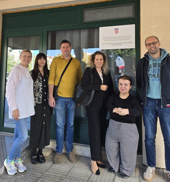 Na fotografiji je šest osoba okupljenih ispred ulaza u zgradu, pozirajući za zajedničku fotografiju. Skupinu čine predstavnici partnerskih organizacija i korisnica projekta, a iza njih na staklenim vratima istaknut je plakat s grbom Republike Hrvatske i informacijom o Programu prekogranične suradnje između Republike Hrvatske i Bosne i Hercegovine. Sudionici stoje nasmiješeni ispred ulaza, dok jedna osoba sjedi u prvom planu. Fotografija je snimljena na otvorenom, pri dnevnom svjetlu, te odaje ozračje suradnje i zajedništva. Istaknuti plakat u pozadini upućuje na provedbu projekta usmjerenog jačanju prekogranične suradnje i unaprjeđenju podrške osobama s invaliditetom i drugim ranjivim skupinama.