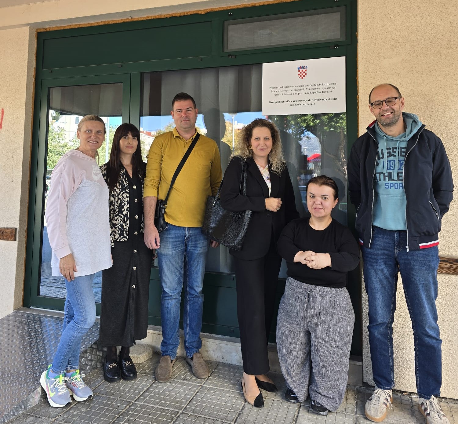 Na fotografiji je šest osoba okupljenih ispred ulaza u zgradu, pozirajući za zajedničku fotografiju. Skupinu čine predstavnici partnerskih organizacija i korisnica projekta, a iza njih na staklenim vratima istaknut je plakat s grbom Republike Hrvatske i informacijom o Programu prekogranične suradnje između Republike Hrvatske i Bosne i Hercegovine. Sudionici stoje nasmiješeni ispred ulaza, dok jedna osoba sjedi u prvom planu. Fotografija je snimljena na otvorenom, pri dnevnom svjetlu, te odaje ozračje suradnje i zajedništva. Istaknuti plakat u pozadini upućuje na provedbu projekta usmjerenog jačanju prekogranične suradnje i unaprjeđenju podrške osobama s invaliditetom i drugim ranjivim skupinama.