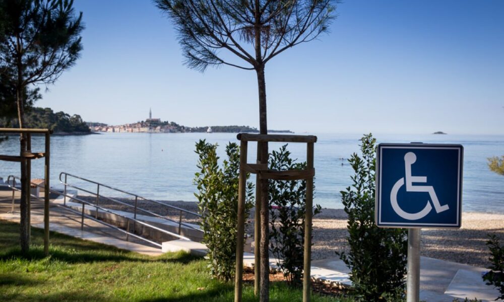 Na fotografiji je prikazan pristupačan dio obale u Rovinju. U prvom planu nalazi se plava oznaka za osobe s invaliditetom, a uz plažu se vidi blaga pristupna rampa s rukohvatima koja omogućuje lakši dolazak do mora osobama u invalidskim kolicima i smanjene pokretljivosti. Uz šetnicu rastu mlada stabla i uređene zelene površine, dok se u pozadini pruža mirno more s pogledom na staru gradsku jezgru Rovinja i prepoznatljiv zvonik crkve sv. Eufemije. Vedro nebo i mirna morska površina dodatno naglašavaju ugodnu i pristupačnu atmosferu ovog dijela obale.