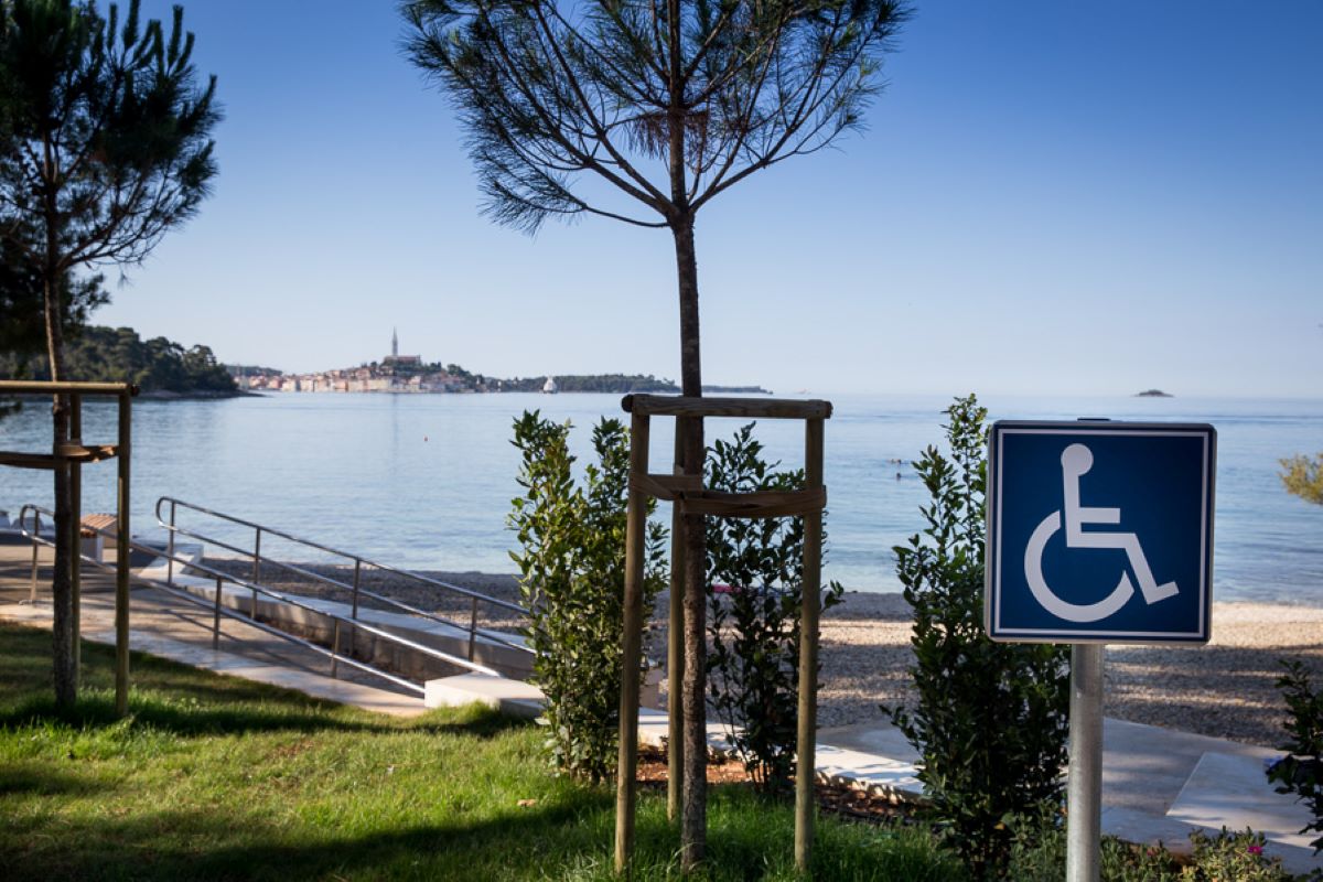 Na fotografiji je prikazan pristupačan dio obale u Rovinju. U prvom planu nalazi se plava oznaka za osobe s invaliditetom, a uz plažu se vidi blaga pristupna rampa s rukohvatima koja omogućuje lakši dolazak do mora osobama u invalidskim kolicima i smanjene pokretljivosti. Uz šetnicu rastu mlada stabla i uređene zelene površine, dok se u pozadini pruža mirno more s pogledom na staru gradsku jezgru Rovinja i prepoznatljiv zvonik crkve sv. Eufemije. Vedro nebo i mirna morska površina dodatno naglašavaju ugodnu i pristupačnu atmosferu ovog dijela obale.