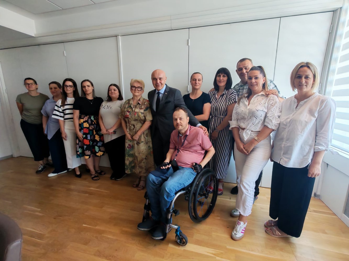 Na fotografiji je snimljena skupina sudionika nakon sastanka u zatvorenom prostoru, vjerojatno u prostorijama Ministarstva. Ukupno je jedanaest osoba koje stoje u polukrugu te jedna osoba u invalidskim kolicima u prvom planu. U sredini skupine nalazi se muškarac u tamnom odijelu i kravati koji pozira zajedno s okupljenima. Sudionici su odjeveni u poslovnu ili poslovno-ležernu odjeću, a svi gledaju prema fotoaparatu i osmjehuju se. Prostorija je svijetla, s drvenim podom, bijelim zidovima i velikim prozorom s desne strane koji propušta prirodno svjetlo. Fotografija odaje dojam službenog susreta i zajedničkog fotografiranja nakon radnog sastanka predstavnika Ministarstva i udruga osoba s invaliditetom.