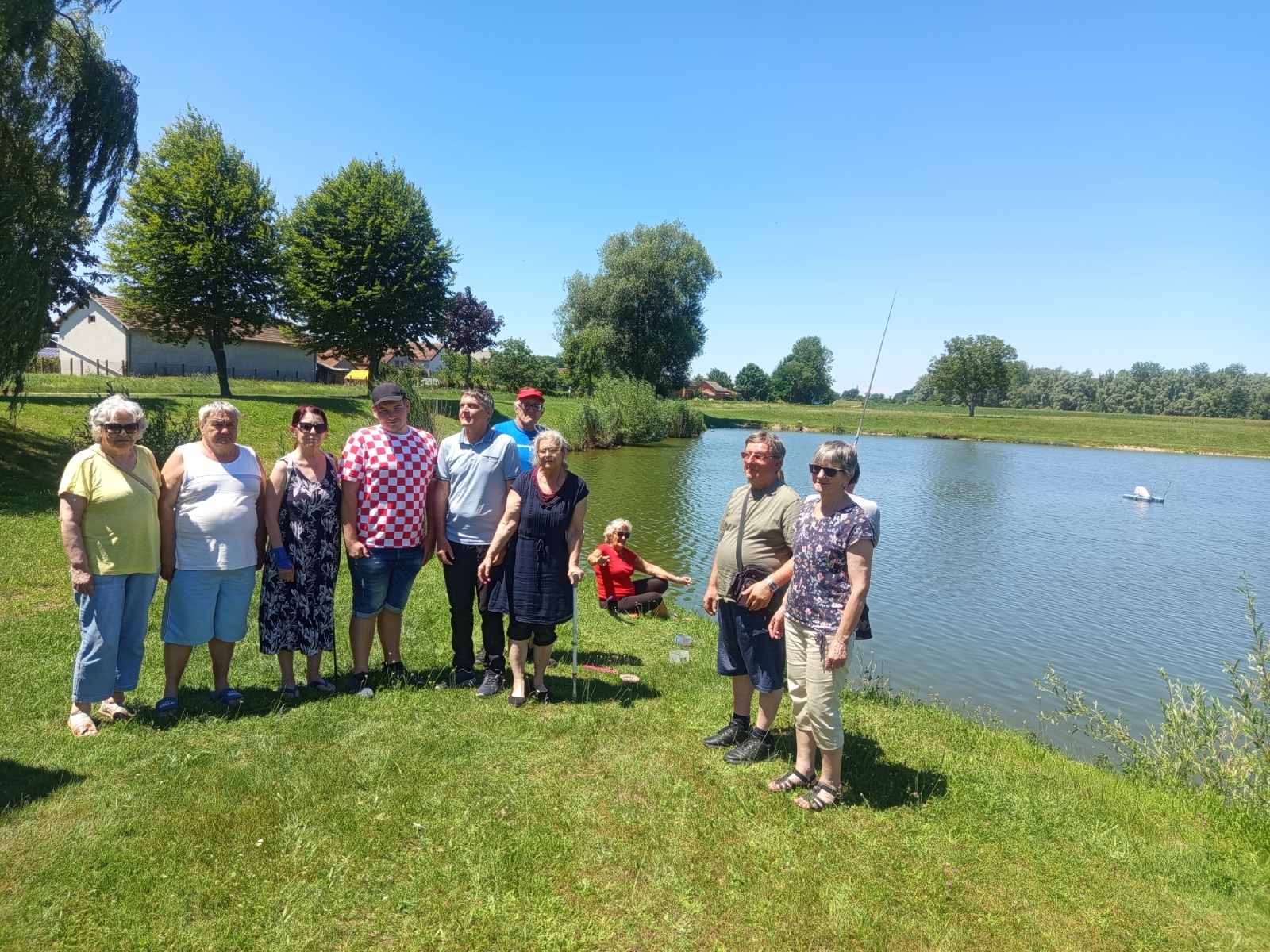 Na fotografiji se nalazi skupina ljudi okupljena uz mirno jezero tijekom sunčanog ljetnog dana. Većina sudionika stoji na uređenoj travnatoj obali i pozira za zajedničku fotografiju, dok jedna osoba sjedi na travi uz samu vodu. U pozadini se vide zelena stabla, livade i nekoliko kuća, što odaje dojam mirnog seoskog krajolika. Vrijeme je vedro, s potpuno plavim nebom, a snažno sunce dodatno naglašava ljetni ugođaj. Na jezeru se uočava mala plutajuća naprava, dok jedan od sudionika u blizini obale drži ribolovni štap. Fotografija odiše opuštenom i prijateljskom atmosferom te prikazuje zajedničko druženje u prirodi.