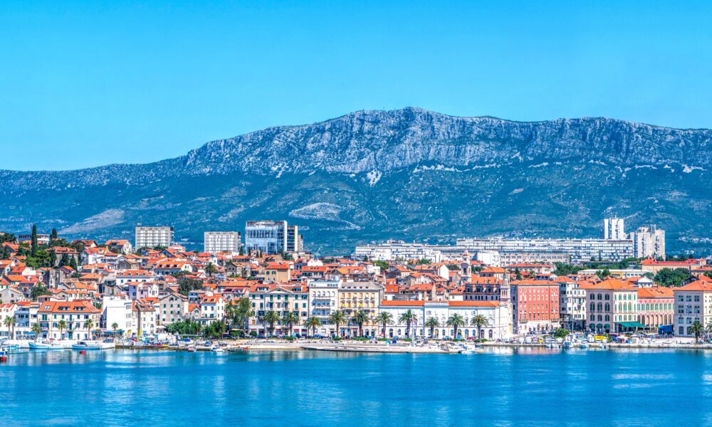 Fotografija prikazuje panoramski pogled na Split s mora. U prvom planu nalazi se mirna, plava morska površina, dok se uz obalu proteže splitska Riva s palmama, brodicama i karakterističnim povijesnim zgradama. U središnjem i donjem dijelu fotografije vidi se stara gradska jezgra Splita, prepoznatljiva po gusto zbijenim kamenim kućama i brojnim krovovima prekrivenima tradicionalnim crvenim crijepom. Uz njih se ističu veće povijesne i moderne građevine, dok se u pozadini pruža urbani dio grada. Iznad Splita dominira planina Mosor, čiji se veliki, kameniti greben proteže cijelom pozadinom. Vedro plavo nebo i intenzivne plave nijanse mora i planine stvaraju vrlo upečatljiv kontrast s toplim crvenim krovovima grada. Fotografija tako prikazuje Split kao spoj povijesne arhitekture, mediteranskog krajolika, mora i planinskog zaleđa, karakterističan za vizuru grada.