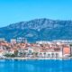 Fotografija prikazuje panoramski pogled na Split s mora. U prvom planu nalazi se mirna, plava morska površina, dok se uz obalu proteže splitska Riva s palmama, brodicama i karakterističnim povijesnim zgradama. U središnjem i donjem dijelu fotografije vidi se stara gradska jezgra Splita, prepoznatljiva po gusto zbijenim kamenim kućama i brojnim krovovima prekrivenima tradicionalnim crvenim crijepom. Uz njih se ističu veće povijesne i moderne građevine, dok se u pozadini pruža urbani dio grada. Iznad Splita dominira planina Mosor, čiji se veliki, kameniti greben proteže cijelom pozadinom. Vedro plavo nebo i intenzivne plave nijanse mora i planine stvaraju vrlo upečatljiv kontrast s toplim crvenim krovovima grada. Fotografija tako prikazuje Split kao spoj povijesne arhitekture, mediteranskog krajolika, mora i planinskog zaleđa, karakterističan za vizuru grada.