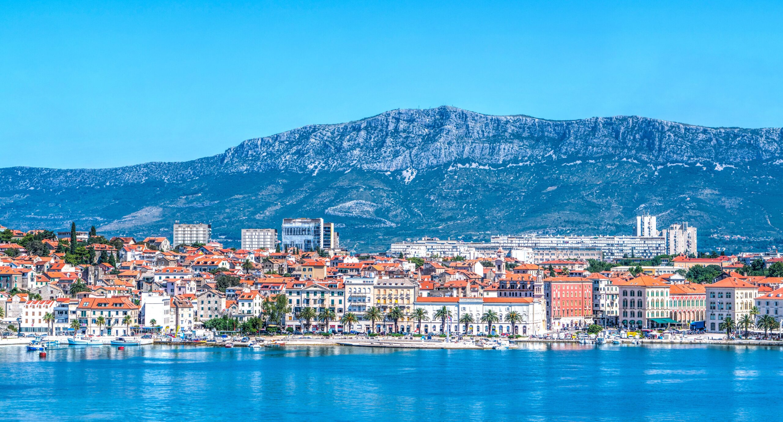Fotografija prikazuje panoramski pogled na Split s mora. U prvom planu nalazi se mirna, plava morska površina, dok se uz obalu proteže splitska Riva s palmama, brodicama i karakterističnim povijesnim zgradama. U središnjem i donjem dijelu fotografije vidi se stara gradska jezgra Splita, prepoznatljiva po gusto zbijenim kamenim kućama i brojnim krovovima prekrivenima tradicionalnim crvenim crijepom. Uz njih se ističu veće povijesne i moderne građevine, dok se u pozadini pruža urbani dio grada. Iznad Splita dominira planina Mosor, čiji se veliki, kameniti greben proteže cijelom pozadinom. Vedro plavo nebo i intenzivne plave nijanse mora i planine stvaraju vrlo upečatljiv kontrast s toplim crvenim krovovima grada. Fotografija tako prikazuje Split kao spoj povijesne arhitekture, mediteranskog krajolika, mora i planinskog zaleđa, karakterističan za vizuru grada.