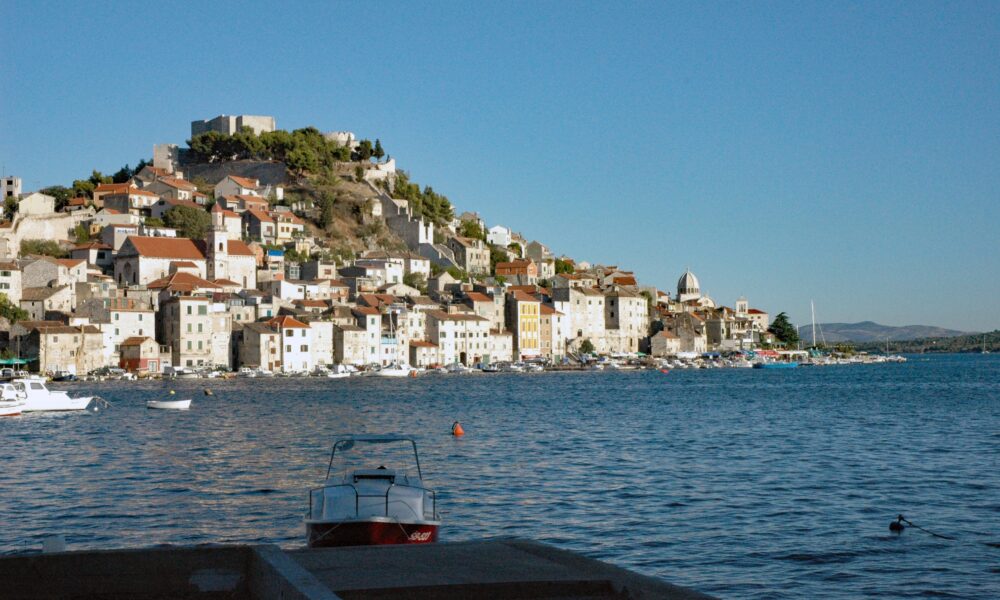 Na fotografiji je panoramski pogled na Šibenik s morske strane, snimljen po vedrom i sunčanom vremenu. U prvom planu nalazi se mirno morsko područje s nekoliko manjih brodica, dok se u pozadini pruža povijesna gradska jezgra. Kuće s karakterističnim crvenim krovovima gusto su smještene na padini, a iznad njih dominira tvrđava sv. Mihovila. U donjem dijelu grada vidljive su kamene kuće uz obalu, brodice i lučki sadržaji. Desno se pruža pogled prema otvorenom moru i okolnom krajoliku. Fotografija posebno ističe mediteranski izgled Šibenika, spoj kamene arhitekture, mora i povijesnih fortifikacija.
