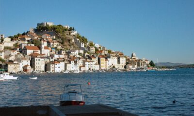 Na fotografiji je panoramski pogled na Šibenik s morske strane, snimljen po vedrom i sunčanom vremenu. U prvom planu nalazi se mirno morsko područje s nekoliko manjih brodica, dok se u pozadini pruža povijesna gradska jezgra. Kuće s karakterističnim crvenim krovovima gusto su smještene na padini, a iznad njih dominira tvrđava sv. Mihovila. U donjem dijelu grada vidljive su kamene kuće uz obalu, brodice i lučki sadržaji. Desno se pruža pogled prema otvorenom moru i okolnom krajoliku. Fotografija posebno ističe mediteranski izgled Šibenika, spoj kamene arhitekture, mora i povijesnih fortifikacija.
