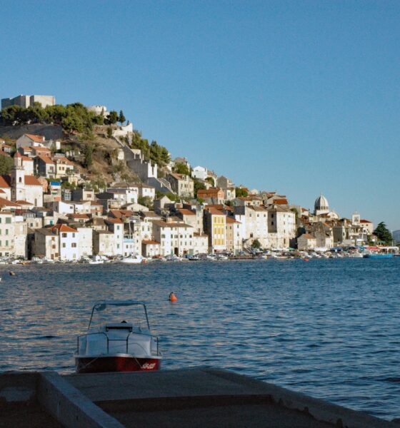 Na fotografiji je panoramski pogled na Šibenik s morske strane, snimljen po vedrom i sunčanom vremenu. U prvom planu nalazi se mirno morsko područje s nekoliko manjih brodica, dok se u pozadini pruža povijesna gradska jezgra. Kuće s karakterističnim crvenim krovovima gusto su smještene na padini, a iznad njih dominira tvrđava sv. Mihovila. U donjem dijelu grada vidljive su kamene kuće uz obalu, brodice i lučki sadržaji. Desno se pruža pogled prema otvorenom moru i okolnom krajoliku. Fotografija posebno ističe mediteranski izgled Šibenika, spoj kamene arhitekture, mora i povijesnih fortifikacija.