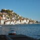 Na fotografiji je panoramski pogled na Šibenik s morske strane, snimljen po vedrom i sunčanom vremenu. U prvom planu nalazi se mirno morsko područje s nekoliko manjih brodica, dok se u pozadini pruža povijesna gradska jezgra. Kuće s karakterističnim crvenim krovovima gusto su smještene na padini, a iznad njih dominira tvrđava sv. Mihovila. U donjem dijelu grada vidljive su kamene kuće uz obalu, brodice i lučki sadržaji. Desno se pruža pogled prema otvorenom moru i okolnom krajoliku. Fotografija posebno ističe mediteranski izgled Šibenika, spoj kamene arhitekture, mora i povijesnih fortifikacija.