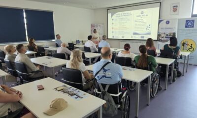 Na fotografiji je prikazana početna konferencija projekta „SRCE ZAJEDNICE“. Sudionici sjede u konferencijskoj dvorani i prate prezentaciju o projektu, dok je jedna osoba ispred projekcijskog platna i predstavlja sadržaj. U prvom planu i među sudionicima vidljive su osobe u invalidskim kolicima, čime fotografija dodatno naglašava uključivost i usmjerenost projekta na osobe s invaliditetom. U pozadini se nalaze promotivni materijali i roll-up plakati projekta „SRCE ZAJEDNICE“.