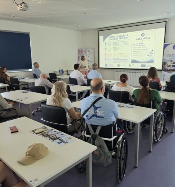 Na fotografiji je prikazana početna konferencija projekta „SRCE ZAJEDNICE“. Sudionici sjede u konferencijskoj dvorani i prate prezentaciju o projektu, dok je jedna osoba ispred projekcijskog platna i predstavlja sadržaj. U prvom planu i među sudionicima vidljive su osobe u invalidskim kolicima, čime fotografija dodatno naglašava uključivost i usmjerenost projekta na osobe s invaliditetom. U pozadini se nalaze promotivni materijali i roll-up plakati projekta „SRCE ZAJEDNICE“.