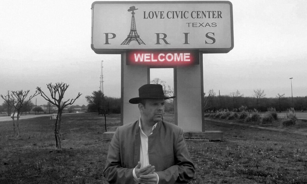 Crno-bijela fotografija muškarca s kaubojskim šeširom ispred znaka ‘Paris’ i ‘Love Civic Center’ u Teksasu.