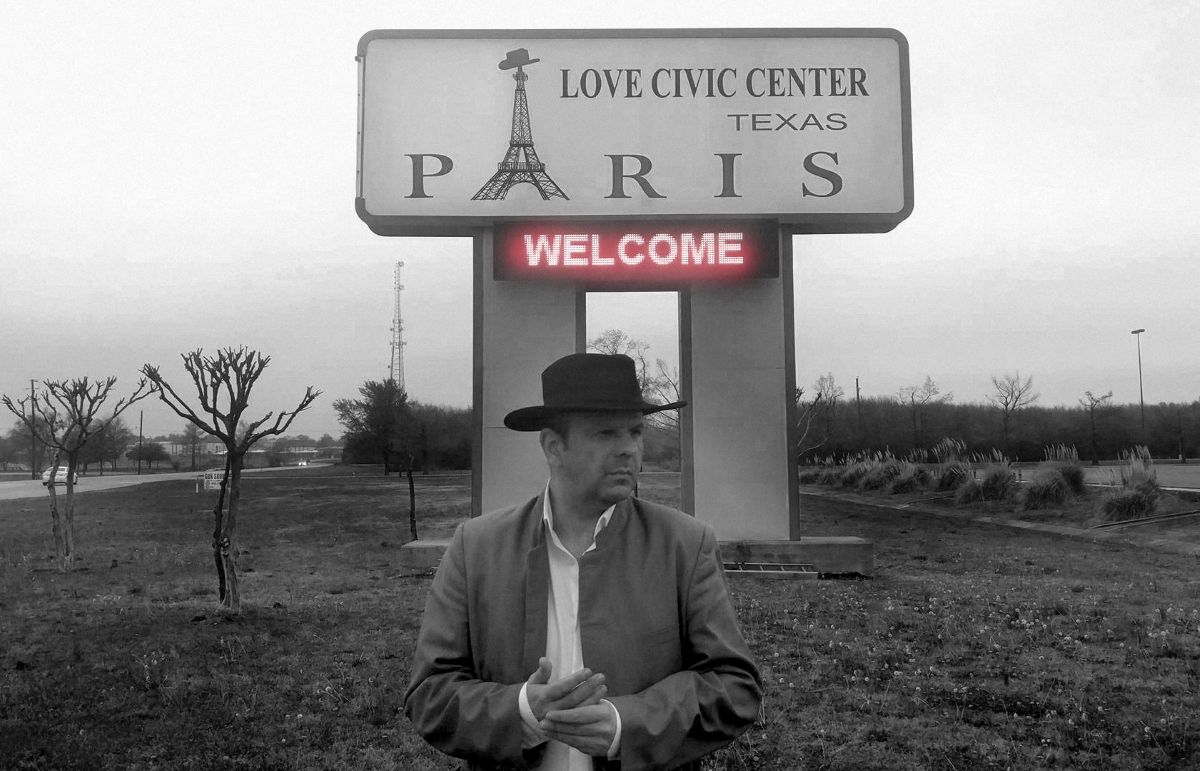 Crno-bijela fotografija muškarca s kaubojskim šeširom ispred znaka ‘Paris’ i ‘Love Civic Center’ u Teksasu.