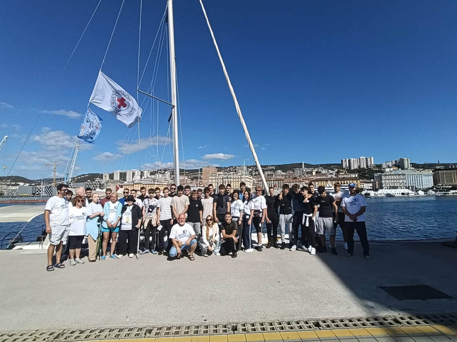 Na fotografiji je velika skupina sudionika projekta „Jedra duha“ okupljena na riječkoj rivi, ispred jedrilice. U prvom planu stoje i sjede sudionici različitih generacija, a među njima su i mladi sudionici te članovi organizacijskog i partnerskog tima. U pozadini se vidi riječki zaljev s gradskom obalom i prepoznatljivom panoramom Rijeke. Na jedrilici se vijore zastave Hrvatskog Crvenog križa i projekta, dok vedro nebo i sunčano vrijeme stvaraju vedru atmosferu. Fotografija dobro prikazuje zajedništvo, inkluziju i druženje sudionika projekta „Jedra duha“, kao i povezanost projekta s morem i jedrenjem.