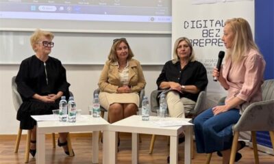 Na slici je prikazan okrugli stol o digitalnoj pristupačnosti. U prostoriji sjede četiri žene koje sudjeluju u razgovoru. Jedna od njih drži mikrofon i govori, dok ostale tri slušaju. U pozadini se nalazi ekran s prikazom online sastanka, a uz njega je vidljiv plakat povezan s temom digitalne pristupačnosti. Ispred sudionica nalaze se stolovi s bocama vode i papirima. Atmosfera djeluje službeno, ali opušteno, a razgovor je usmjeren na razmjenu mišljenja i iskustava.