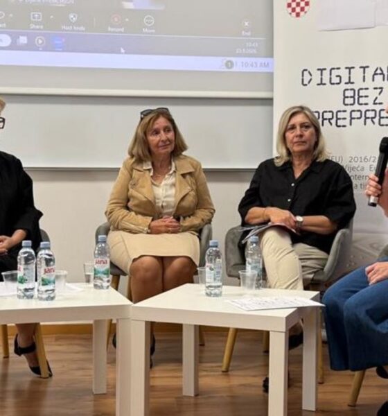 Na slici je prikazan okrugli stol o digitalnoj pristupačnosti. U prostoriji sjede četiri žene koje sudjeluju u razgovoru. Jedna od njih drži mikrofon i govori, dok ostale tri slušaju. U pozadini se nalazi ekran s prikazom online sastanka, a uz njega je vidljiv plakat povezan s temom digitalne pristupačnosti. Ispred sudionica nalaze se stolovi s bocama vode i papirima. Atmosfera djeluje službeno, ali opušteno, a razgovor je usmjeren na razmjenu mišljenja i iskustava.