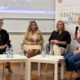 Na slici je prikazan okrugli stol o digitalnoj pristupačnosti. U prostoriji sjede četiri žene koje sudjeluju u razgovoru. Jedna od njih drži mikrofon i govori, dok ostale tri slušaju. U pozadini se nalazi ekran s prikazom online sastanka, a uz njega je vidljiv plakat povezan s temom digitalne pristupačnosti. Ispred sudionica nalaze se stolovi s bocama vode i papirima. Atmosfera djeluje službeno, ali opušteno, a razgovor je usmjeren na razmjenu mišljenja i iskustava.