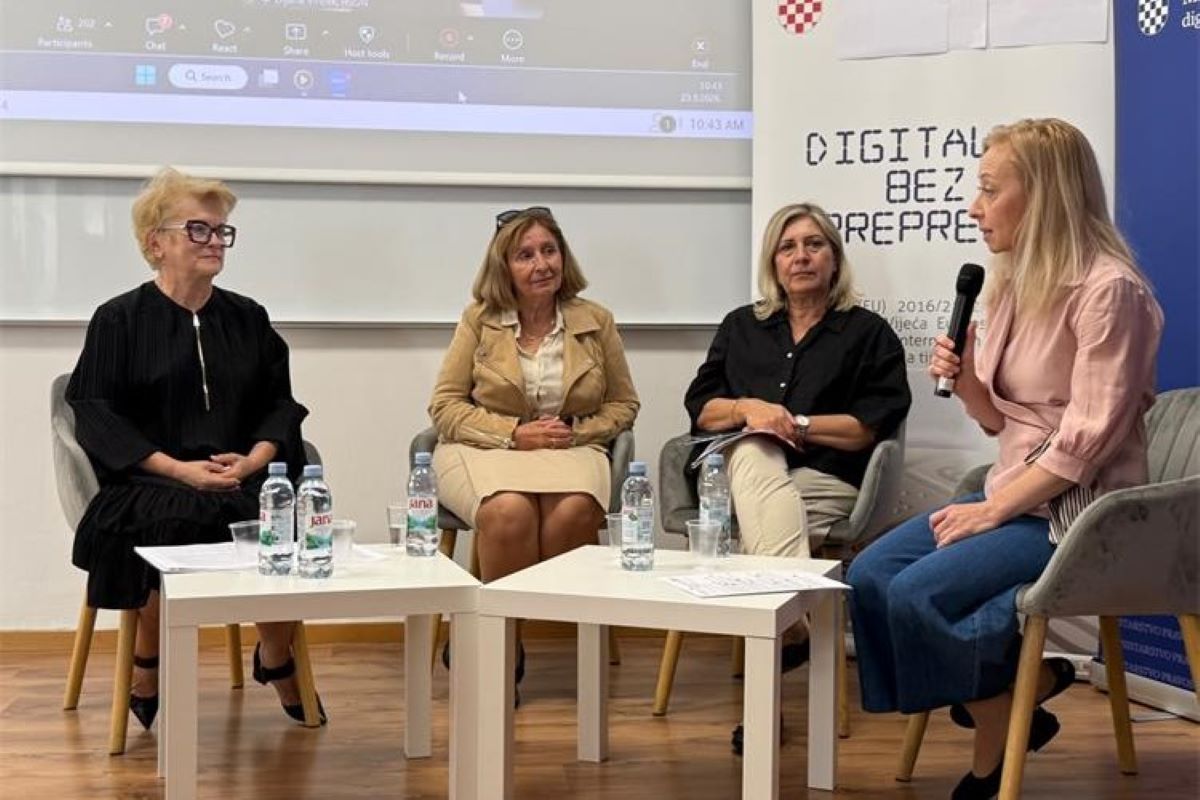 Na slici je prikazan okrugli stol o digitalnoj pristupačnosti. U prostoriji sjede četiri žene koje sudjeluju u razgovoru. Jedna od njih drži mikrofon i govori, dok ostale tri slušaju. U pozadini se nalazi ekran s prikazom online sastanka, a uz njega je vidljiv plakat povezan s temom digitalne pristupačnosti. Ispred sudionica nalaze se stolovi s bocama vode i papirima. Atmosfera djeluje službeno, ali opušteno, a razgovor je usmjeren na razmjenu mišljenja i iskustava.
