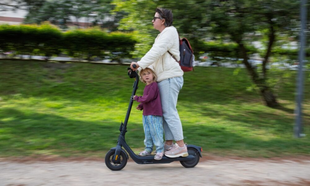Odrasla osoba i dijete zajedno se voze na električnom romobilu kroz park.