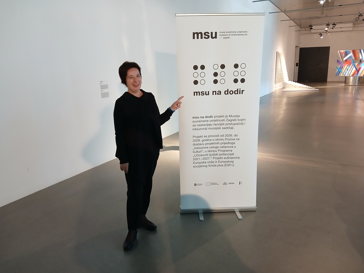 Žena u crnoj odjeći pokazuje na informativni pano projekta „MSU na dodir“ u Muzeju suvremene umjetnosti u Zagrebu.