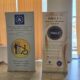 Na fotografiji se nalaze dva promotivna roll-up plakata Udruge osoba s invaliditetom Grada Požege i Požeško-slavonske županije. Na desnom plakatu predstavljen je program „DiKA 2 – Društvene i kreativne aktivnosti“, dok se na lijevom nalazi simbol pristupačnosti i ilustracija koja prikazuje pružanje pomoći osobi u invalidskim kolicima. Plakati su postavljeni u zatvorenom prostoru, uz prozore prekrivene žutim zavjesama. Fotografija prikazuje promotivne materijale kojima se predstavljaju rad Udruge, program DiKA 2 i važnost podrške osobama s invaliditetom.