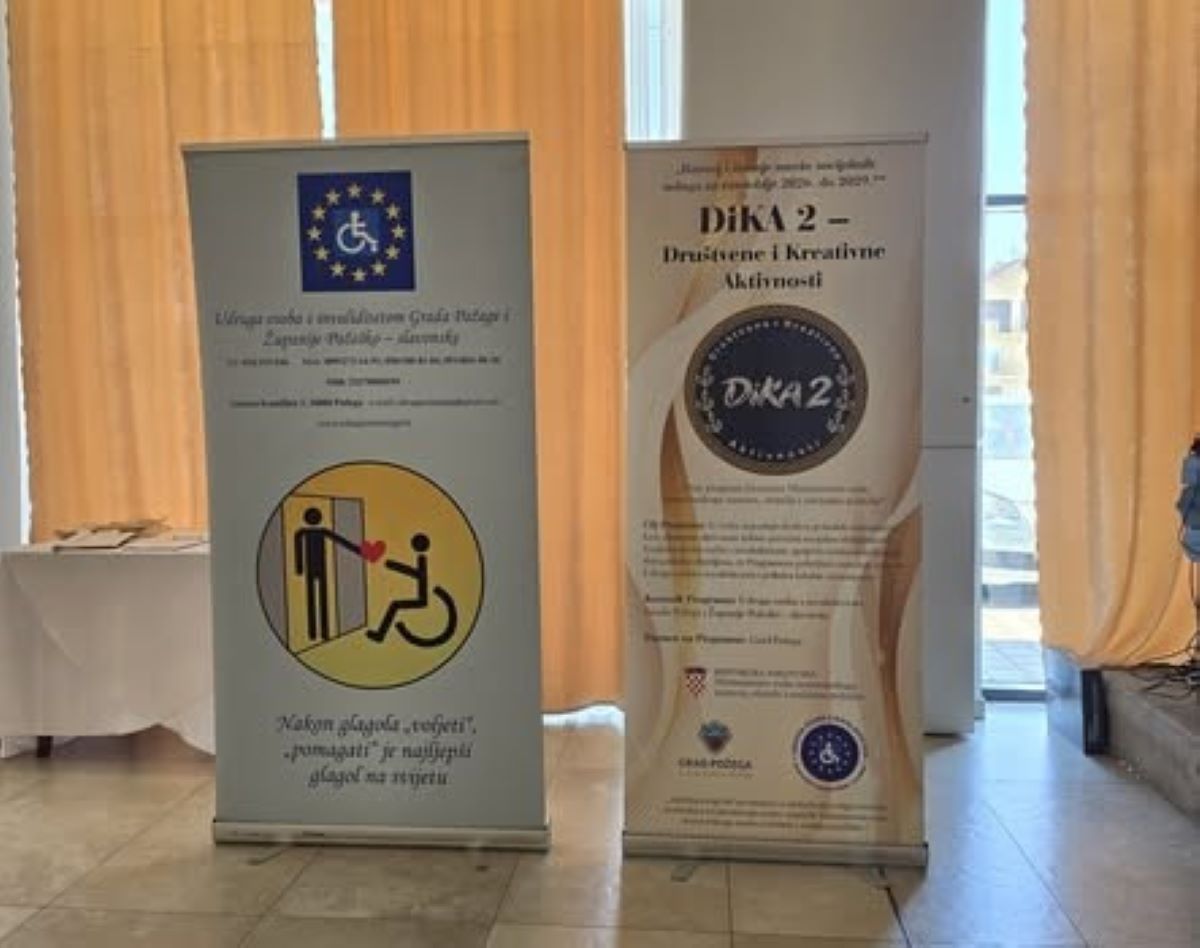 Na fotografiji se nalaze dva promotivna roll-up plakata Udruge osoba s invaliditetom Grada Požege i Požeško-slavonske županije. Na desnom plakatu predstavljen je program „DiKA 2 – Društvene i kreativne aktivnosti“, dok se na lijevom nalazi simbol pristupačnosti i ilustracija koja prikazuje pružanje pomoći osobi u invalidskim kolicima. Plakati su postavljeni u zatvorenom prostoru, uz prozore prekrivene žutim zavjesama. Fotografija prikazuje promotivne materijale kojima se predstavljaju rad Udruge, program DiKA 2 i važnost podrške osobama s invaliditetom.