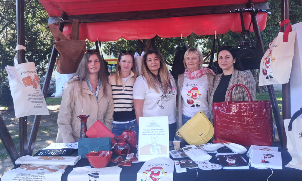 Pet žena pozira iza štanda URIHO-a s izloženim torbama, keramikom i promotivnim proizvodima.