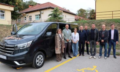 Na fotografiji je novo kombi vozilo Udruge Neven, uz koje pozira skupina predstavnika i suradnika udruge. U pozadini se nalaze zgrade, a na tlu je označeno parkirno mjesto za osobe s invaliditetom. Fotografija prikazuje važan trenutak za udrugu, koja je povodom 53. obljetnice dobila prilagođeno vozilo za prijevoz svojih korisnika.