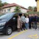 Na fotografiji je novo kombi vozilo Udruge Neven, uz koje pozira skupina predstavnika i suradnika udruge. U pozadini se nalaze zgrade, a na tlu je označeno parkirno mjesto za osobe s invaliditetom. Fotografija prikazuje važan trenutak za udrugu, koja je povodom 53. obljetnice dobila prilagođeno vozilo za prijevoz svojih korisnika.