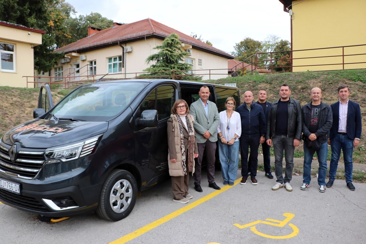 Na fotografiji je novo kombi vozilo Udruge Neven, uz koje pozira skupina predstavnika i suradnika udruge. U pozadini se nalaze zgrade, a na tlu je označeno parkirno mjesto za osobe s invaliditetom. Fotografija prikazuje važan trenutak za udrugu, koja je povodom 53. obljetnice dobila prilagođeno vozilo za prijevoz svojih korisnika.