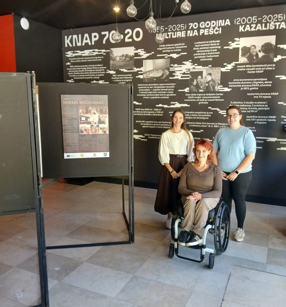 Tri žene poziraju ispred izložbenog panoa o 70 godina Kazališta KNAP, među njima žena u invalidskim kolicima.