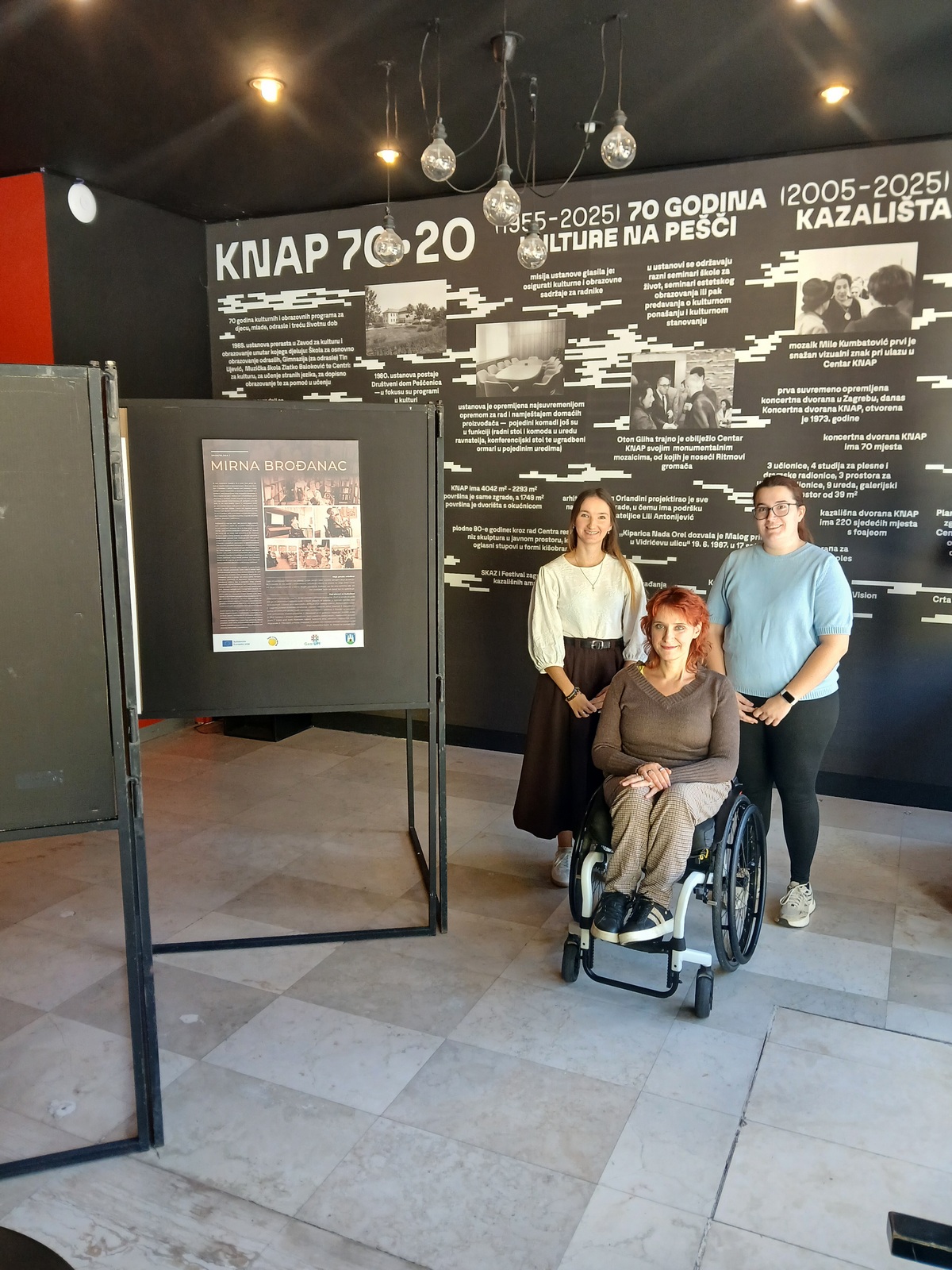 Tri žene poziraju ispred izložbenog panoa o 70 godina Kazališta KNAP, među njima žena u invalidskim kolicima.