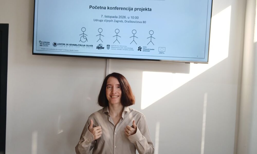 Žena pokazuje palčeve gore ispred zaslona s najavom projekta „Da ti pokažem što mogu?“ Udruge slijepih Zagreb.