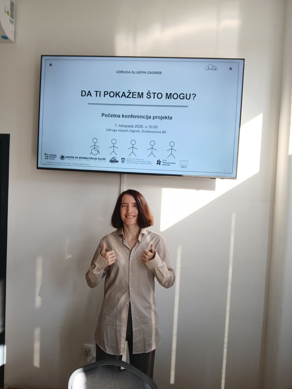Žena pokazuje palčeve gore ispred zaslona s najavom projekta „Da ti pokažem što mogu?“ Udruge slijepih Zagreb.