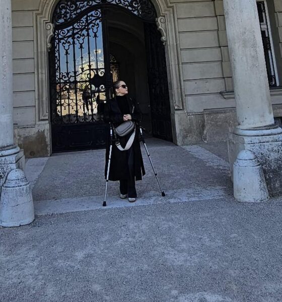 Žena u crnom kaputu i sa štakama stoji ispred ukrašenih željeznih vrata povijesne zgrade.