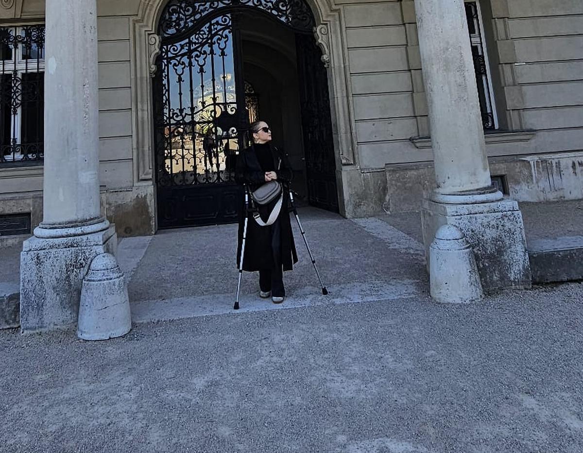 Žena u crnom kaputu i sa štakama stoji ispred ukrašenih željeznih vrata povijesne zgrade.