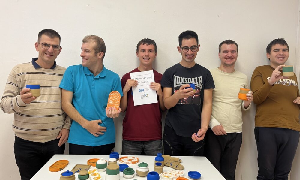Na fotografiji je šest sudionika projekta „Manufaktura dobrote” koji s ponosom predstavljaju predmete izrađene na kreativnim radionicama. Na stolu ispred njih izložene su šarene posudice, svijećnjaci i ukrasni predmeti različitih oblika i boja. Fotografija prikazuje zajednički rad, kreativnost i zadovoljstvo sudionika postignutim rezultatima.