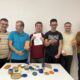 Na fotografiji je šest sudionika projekta „Manufaktura dobrote” koji s ponosom predstavljaju predmete izrađene na kreativnim radionicama. Na stolu ispred njih izložene su šarene posudice, svijećnjaci i ukrasni predmeti različitih oblika i boja. Fotografija prikazuje zajednički rad, kreativnost i zadovoljstvo sudionika postignutim rezultatima.