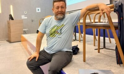 Muškarac s bradom sjedi na rubu pozornice u konferencijskoj dvorani, oslonjen rukom na naslon stolice.
