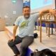 Muškarac s bradom sjedi na rubu pozornice u konferencijskoj dvorani, oslonjen rukom na naslon stolice.
