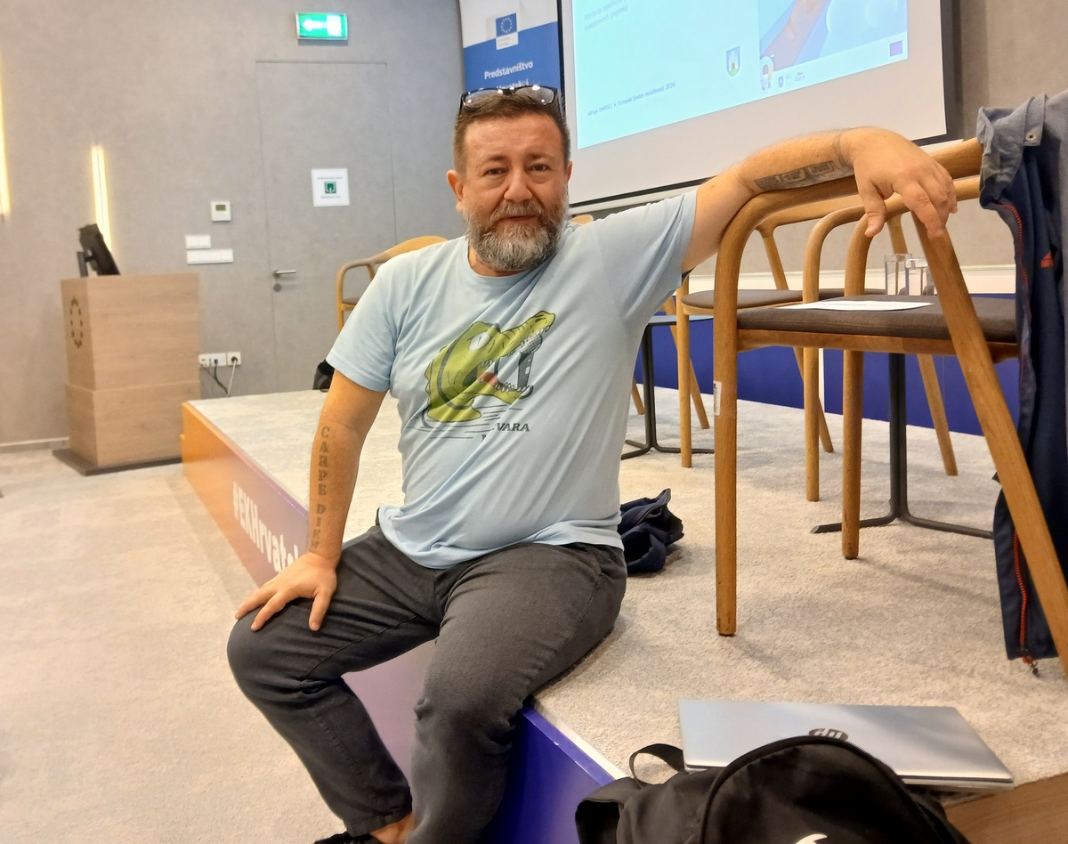 Muškarac s bradom sjedi na rubu pozornice u konferencijskoj dvorani, oslonjen rukom na naslon stolice.